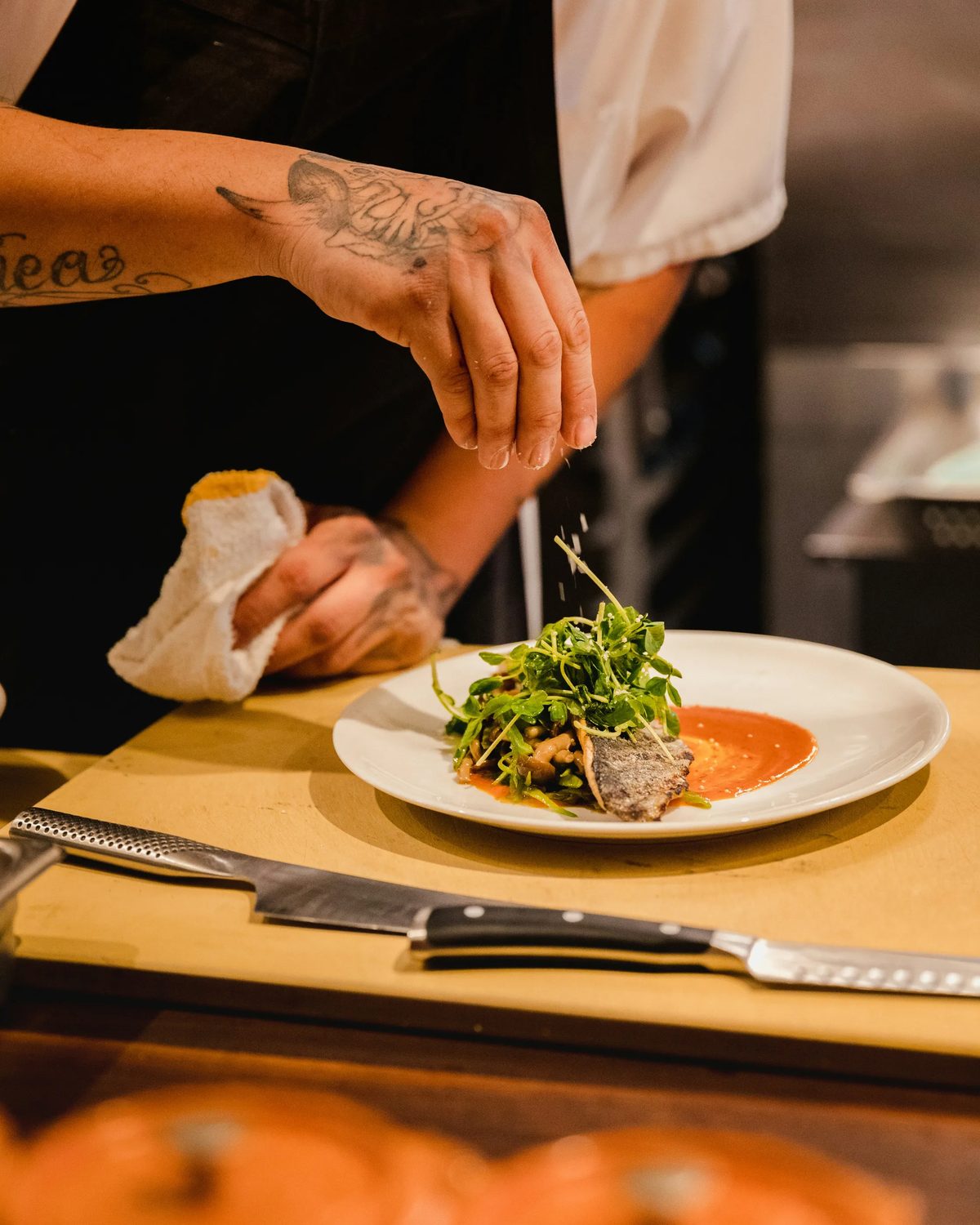 A chef seasoning a plate of fish and greens at the pass